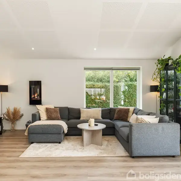 En grå hjørnesofa står i en stue, omgivet af grønne planter, et rundt sofabord og en stor fladskærms-tv på sort møbel. Vinduet slipper lys ind.