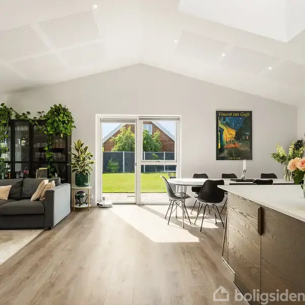 Stue med sofa og rundt bord til venstre, spisestuebord i midten, køkkenø med blomster til højre. Store vinduer og glasdør giver udsigt til solrig have.