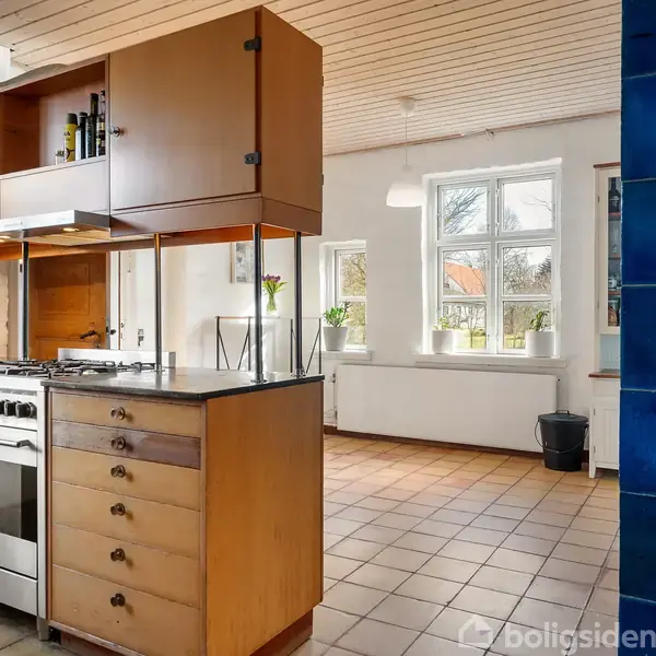 Et køkken med en fritstående komfurø og træskabe. Lyse klinker på gulvet, et vindue med udsigt udenfor og en blå flisevæg i højre hjørne.
