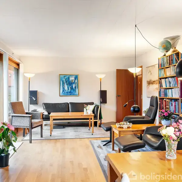 Stue med sofa og lænestole ved sofabord, omgivet af reol fyldt med bøger. Store vinduer til venstre med udsigt mod have. Blomster på et bord.