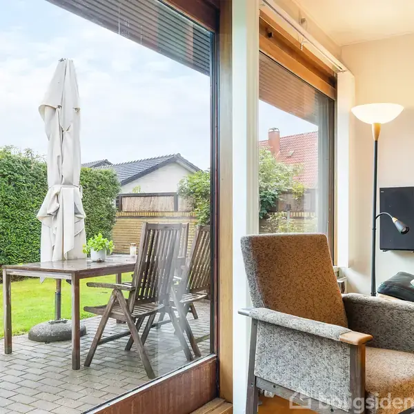 Indendørs stol placeret nær et glasvindue med udsigt til en terrasse. Udendørs står et bord med stole og en sammenfoldet parasol. Haven er omgivet af en hæk.
