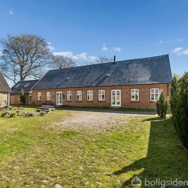 Et rødt murstenshus med sorte tagsten står stille i en åbning mellem to udhuse. Græsplæne og stenbelagt gårdsplads omgiver huset, med træer i baggrunden under en blå himmel.