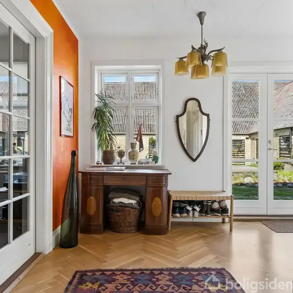Entré med træbord dekoreret med planter og flasker, væghængt spejl. Trædøre med glas åbner til haveområde. Orange væg og tæppe tilføjer farve ved siden af rummet med blå sofa.