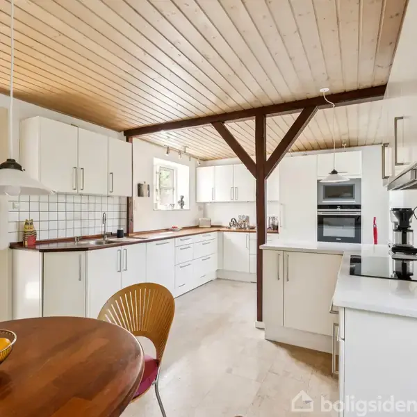 Et lyst køkken med hvide skabe og træloft har en træbænk med skåle fyldt med appelsiner. En stol står ved bordet, mens moderne apparater er indbygget i skabene.