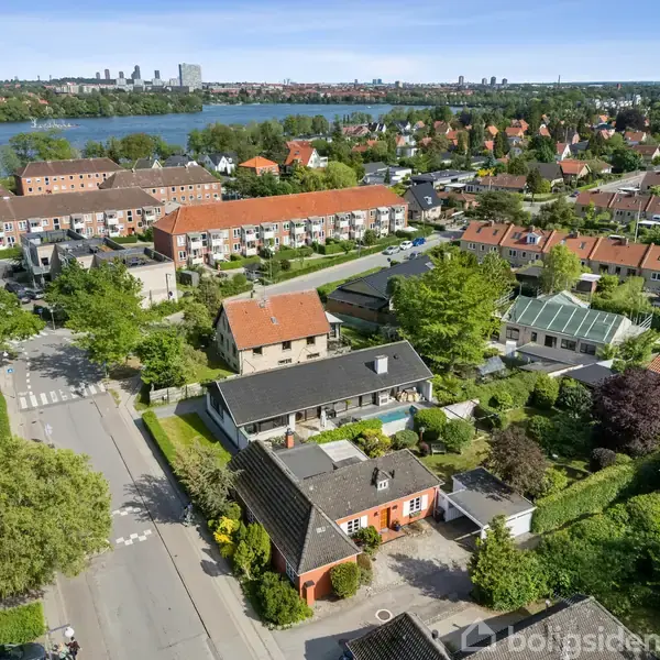 En række huse ligger stille i et grønt boligområde med træer og haver. I baggrunden ses en sø og flere boligblokke i en bymæssig setting.