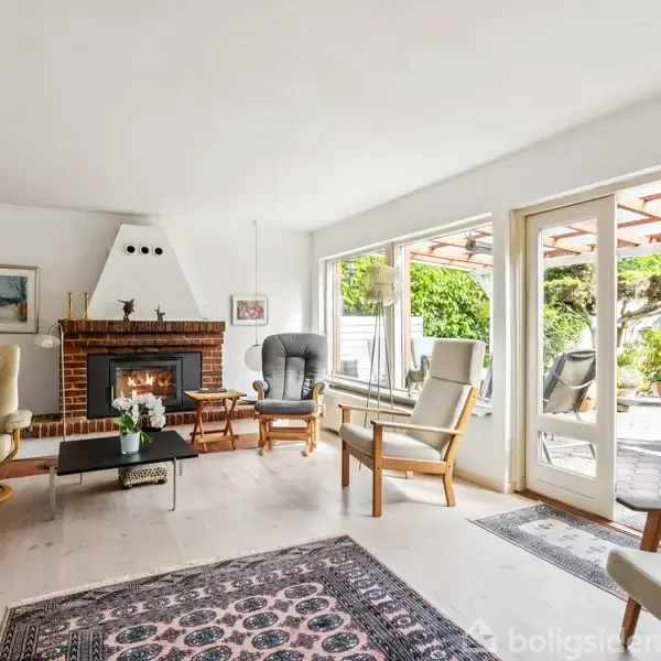 Stue med pejs, hvor stole og borde er placeret omkring den. Store vinduer giver udsigt til en terrasse med planter. Lyse trægulve og en tæppe dekorerer rummet.