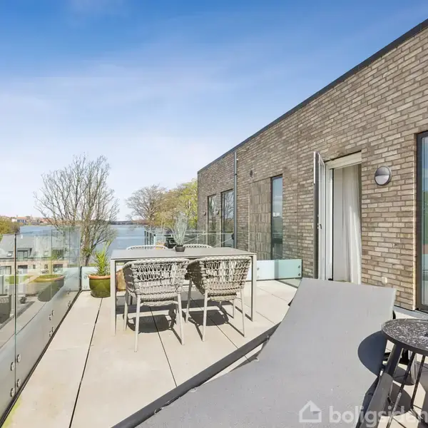 Terrasse med liggestole og et bord, placeret foran en murstensbygning. Et spisebord med stole står ved siden af, omgivet af glasrækværk. Træer og hustage kan ses i baggrunden.