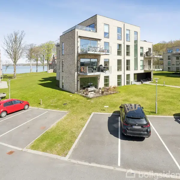 En moderne lejlighedsbygning med store vinduer står omgivet af græsplæner og parkerede biler. I baggrunden ses et landskab med træer og vand.