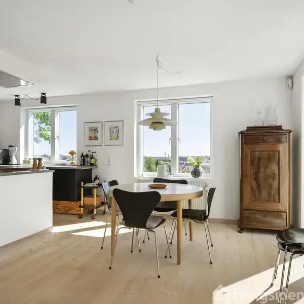Et stilfuldt køkken-alrum viser et rundt bord med sorte stole, ved siden af en pejs. Et enkelt vitrineskab står i hjørnet. Sollys strømmer ind fra store vinduer.