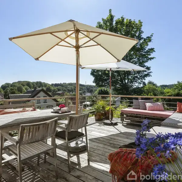 Terrasse med træmøbler under store parasoller, der skaber skygge. Omkring er en frodig udsigt med træer og hustage i baggrunden. Blomster og puder tilføjer farve og hygge.