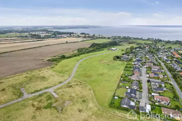 Landskab set ovenfra, viser et boligkvarter med rækkehuse tæt ved åbne marker og en kystlinje i baggrunden. Vejnet forbinder området, omgivet af grønne marker og havudsigt.