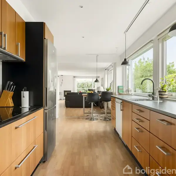 Køkken med træskabe og en sølvfarvet køleskab, står ubrugt. Store vinduer og træbordplade, lysner rummet, som fører til en spise- og stuearea. Lyst og moderne interiør.