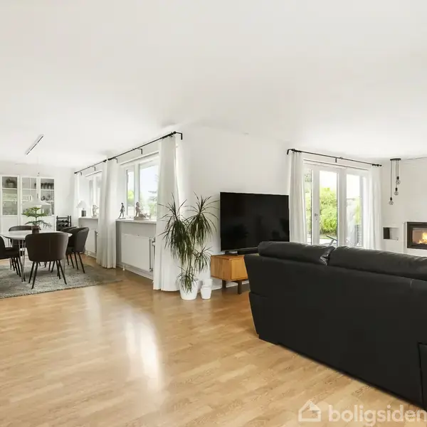 Sort lædersofa står i en lys stue med trægulv, vendt mod en stor fladskærms-tv. Ved siden af er spisebord med stole; et vindue bagved åbner ud til en frodig have.