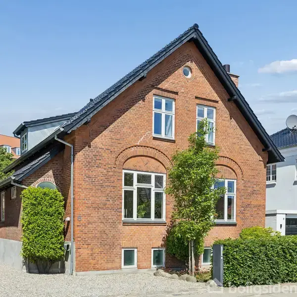 Et rødt murstenshus med sort tag står i en grusbelagt indkørsel. Huset har store vinduer, og grønne buske omkranser indgangen. Det er en solrig dag med blå himmel og få skyer.