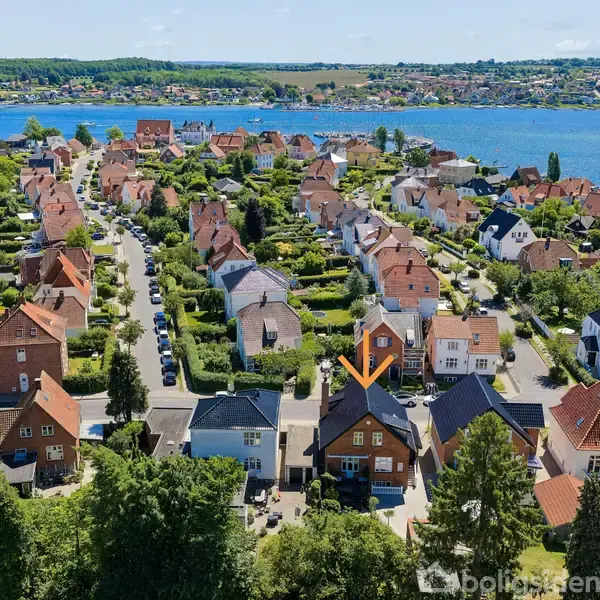 Luftfoto af et boligområde med taghuse. En orange pil peger på et specifikt hus. Området er tæt bebygget, omgivet af grønne områder og ligger ved en sø.