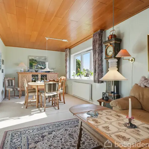 En stue med sofa og sofabord i forgrunden. Et spisebord med stole står nær en væg med billeder. Vindue og bordlampe tilføjer lys. Tæppe på gulvet.