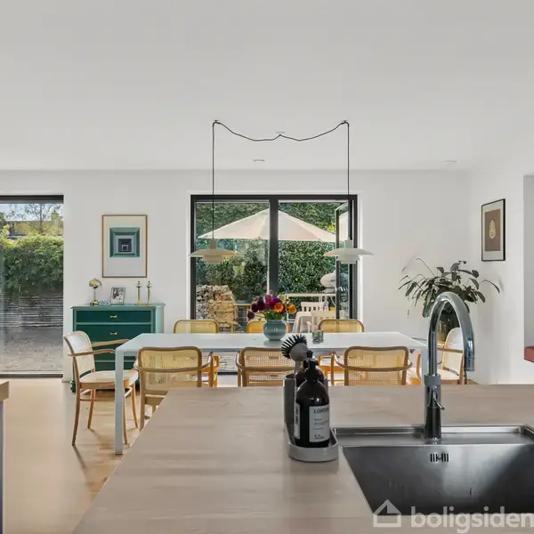 Et moderne køkken-alrum med vask på køkkenø, omgivet af et spisebord med stole. Stort vindue åbner mod en terrasse med parasol. Lyse vægge og grønne planter skaber en frisk atmosfære.