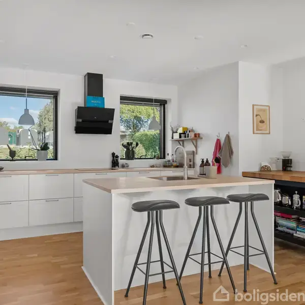 Køkken med tre barstole omkring en central ø, hvide skabe, og vinduer med udsigt til grønt. Hylder med bøger og planter i et lyst rum med trægulv.