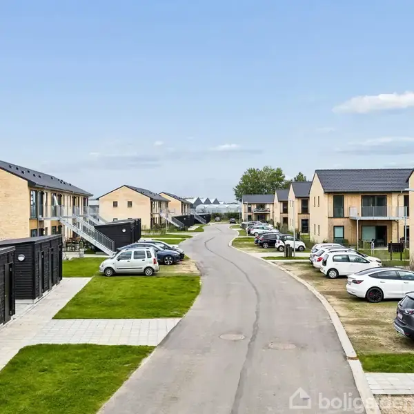 Rækkehuse står parallelt langs en asfalteret vej med grønne plæner og parkerede biler på begge sider, under en klar blå himmel.