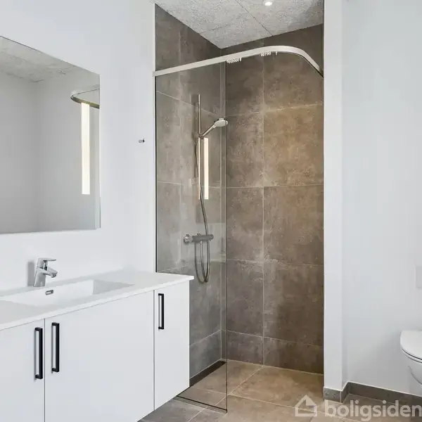 Badeværelse med hvidt vaskeskab, spejl og brusekabine med regnbruser til højre. Vaskemaskine og tørretumbler er placeret til venstre. En væghængt toilet ses ved siden af.