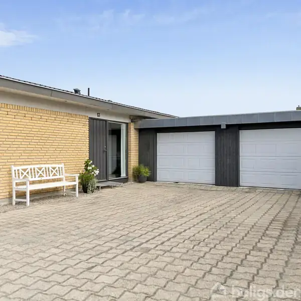 En gulstensbygning med fladt tag står stille ved siden af en flisebelagt gårdsplads. Til venstre er en hvid bænk, mens to hvid-grå garageporte ses til højre.