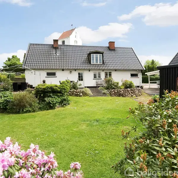 Hvidt hus med mørkt tag står stille i en velholdt have. Blomstrende buske og græsplæne omgiver huset. Skyfri himmel og få skyer danner baggrunden.