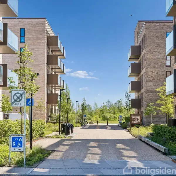 Boligblokke med altaner står langs en gangsti, omgivet af grønne træer og buske. Trafikskilte viser "Zone 4" og "Zone 2". Klar blå himmel i baggrunden.