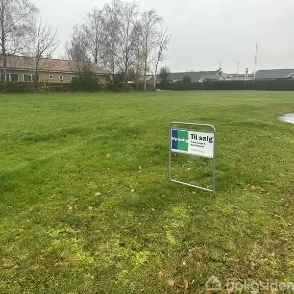 Et græsareal med et "Til salg" skilt fra Nybolig står i forgrunden. I baggrunden ses nogle huse og træer under en grå himmel.