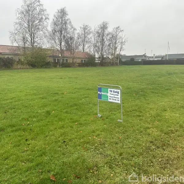 Et skilt står på en grøn græsplæne og viser teksten "Til Salg Nybolig" med en telefonnummer. I baggrunden ses træer og huse under en overskyet himmel.