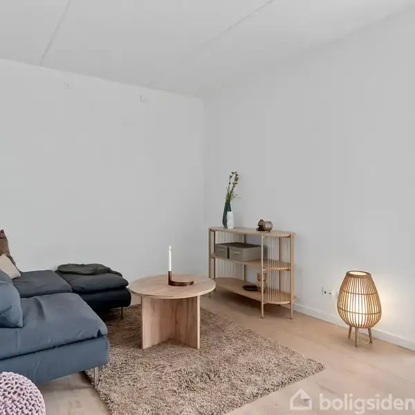 En mørkeblå sofa står i en stue med lyse trægulve. Et lille rundt bord med stearinlys står foran sofaen. Rummet har en reol, gulvlampe og et stort vindue.