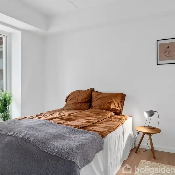 En seng med brune puder og et gråt tæppe står i et lyst soveværelse. Ved siden af sengen er et lille bord med en lampe. En plante står nær vinduet.