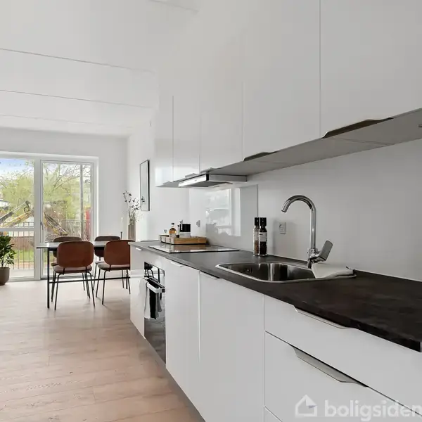 Et moderne køkken med hvide skabe og sort bordplade, hvor vandhanen og kogezonen tilfører funktionalitet. I baggrunden ses spiseplads med bord, stole og adgang til terrasse gennem glasdøre.