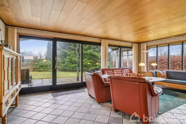 Stue med brune læderstole og glasbord står på klinkegulv. Store vinduer giver udsigt til en grøn have med buske og træer. Træloftet skaber en varm atmosfære.