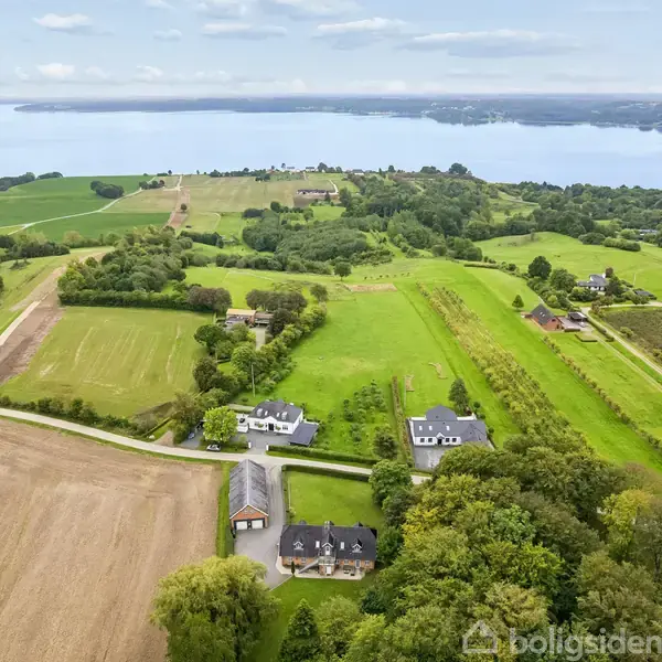 Landområde med huse omgivet af grønne marker og skove, set fra luften. En sø kan ses i baggrunden med en klar himmel ovenover.