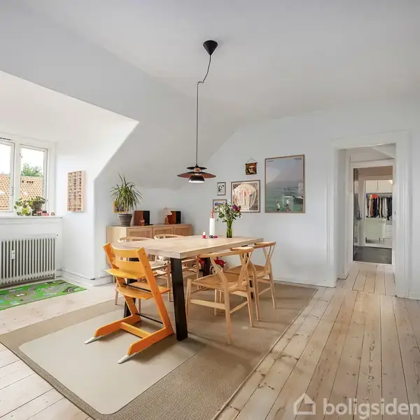 En spisebord med seks stole står i et lyst rum med trægulv. Vindueskarmen har grønne planter. En bogreol og kunstværker pynter væggene.