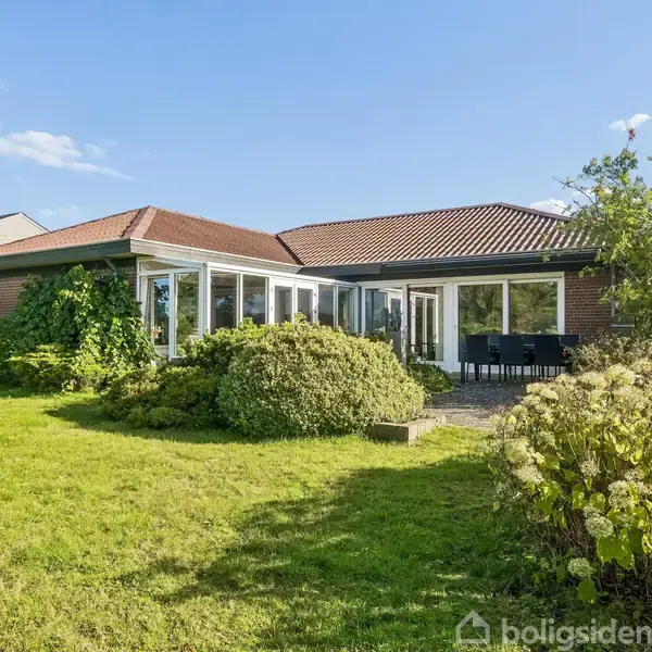 Huset er en etplans villa med stort vinduesparti og rødt tegltag. Det står i en velplejet have med græsplæne, buske og en terrasse med havemøbler under blå himmel.