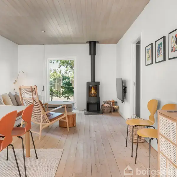 En stue med moderne træmøbler og orange stole omkring et spisebord. Brændeovn i hjørnet. Lyst trægulv, hvidmalede vægge, kunst på væggene og udsigt til have gennem et vindue.