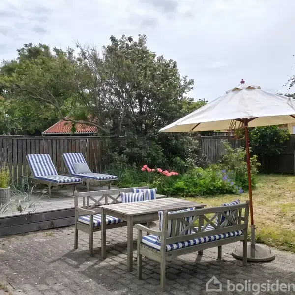 Terrassemøbler står på en flisebelagt patio med to liggestole i baggrunden. En stor paraply dækker et træbord og bænke. Omgivet af græsplæne og træer, indhegnet af et hegn.