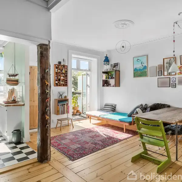 Stue med spisebord og grøn stol, dekoreret med kunst på væggen. Sollys strømmer ind fra et vindue. Ved siden af er et køkken med sort-hvidt ternet gulv.