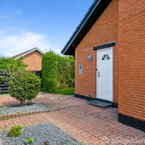 Et murstenshus med en hvid dør står i en have med fliser og grus. Der er buske, et hegn og en anden bygning i baggrunden.