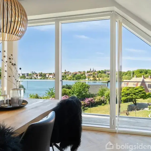 En spisebord med dekorationer står i et lyst rum foran store vinduer. Udsigt over en sø med grønne haver og huse i baggrunden skaber en lys atmosfære.