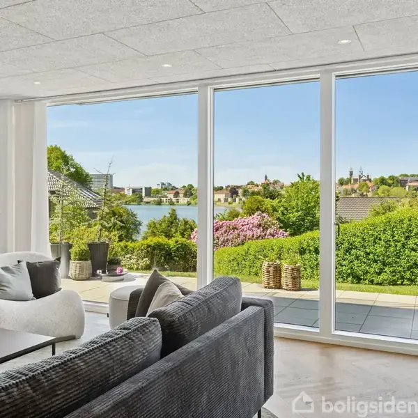 En moderne stue med grå sofaer og et bord, der står vidt ud mod store vinduer. Vinduerne viser en have med grønne buske og udsigt over en sø og by.