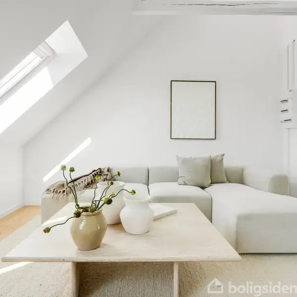 En lys stue med en cremefarvet hjørnesofa står ved en skråtagvæg. Et minimalistisk billede hænger over sofaen. Et lavt bord med vaser og en plante står i forgrunden.