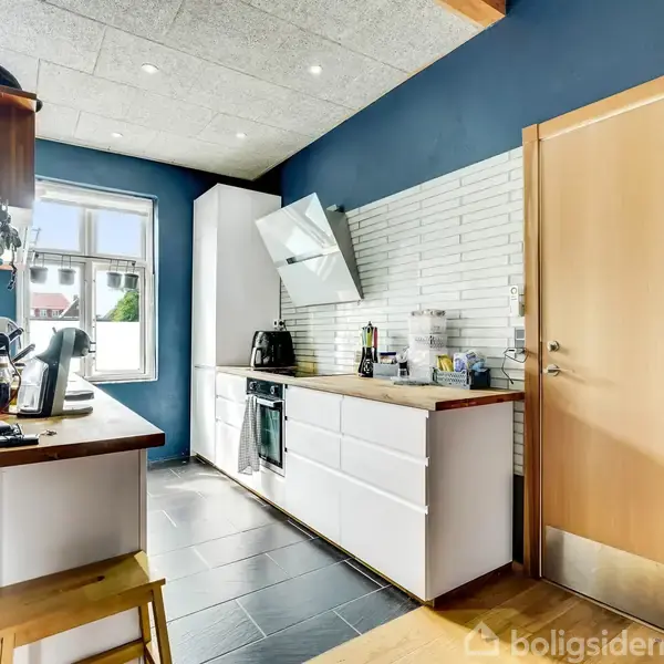 Et moderne køkken med hvide skabe og en sort ovn, hvor en kaffemaskine står på bordet. Rummet har blå vægge, en trædør og et vindue, der giver naturligt lys.