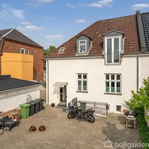 En hvid bygning med rødt tag står i en gårdsplads. En motorcykel er parkeret ved siden af containere og planter. En murstensbygning med oranger facade ses i baggrunden.