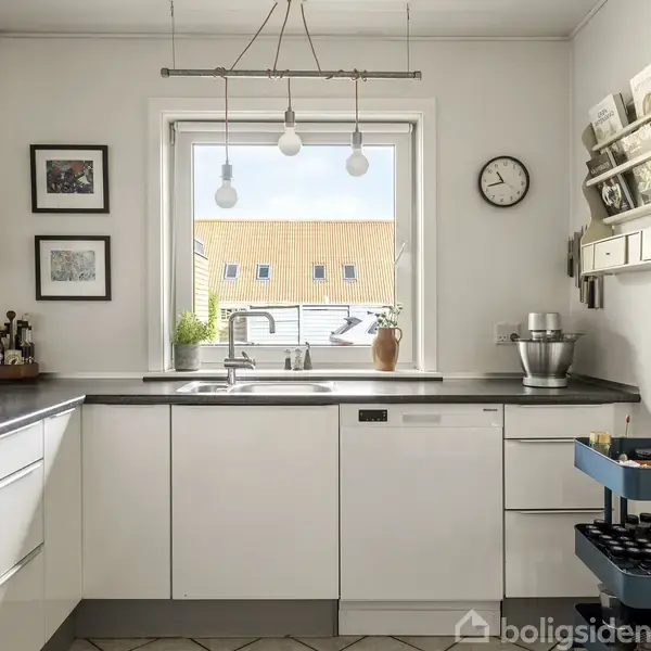 Køkken med hvide skabe i en L-form, hvor en køkkenvask er monteret under et vindue. Ovenfor hænger pendellamper. Vægge pyntet med billeder, hylder, og en vægur.