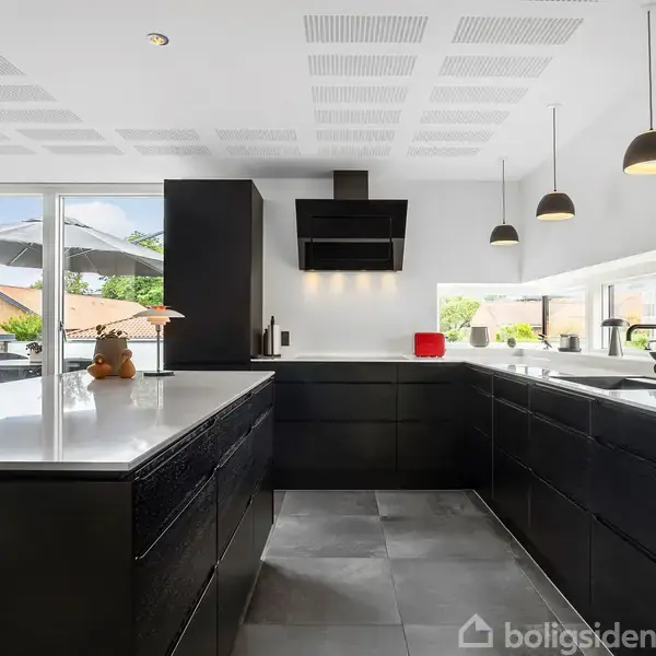Køkken med sorte skabe og hvide bordplader. Tre hængelamper over øen, store vinduer mod en terrasse med parasol. Rummeligt og moderne design med en indbygget ovn.