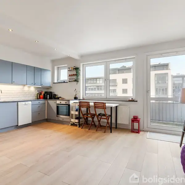 Køkken-alrum med lyse trægulve domineret af et moderne, minimalistisk køkken med blå skabslåger mod venstre. Midt i rummet står et lille bord med to stole ved store vinduer, hvorigennem sollys strømmer ind. Hylder med planter og dekorationer hænger på højre væg.