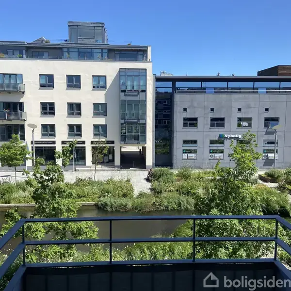 En moderne bygning står ved siden af en vej. Bygningen har store vinduer. I forgrunden er der en balkon med udsigt over en grøn park og vandløb. Skilte: "Nybolig" og "Nykredit".