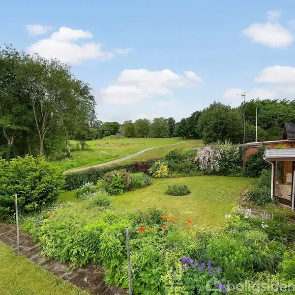 En grøn have fyldt med blomster og buske omkranser en sti, der fører til åbne marker. Ved siden af er der et hus med en terrasse.